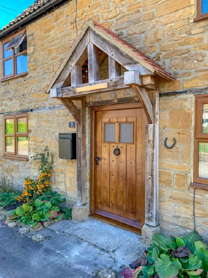 Framed plank oak door with two small glazed lights and a ring knocker beneath an oak porch on a stone cottage