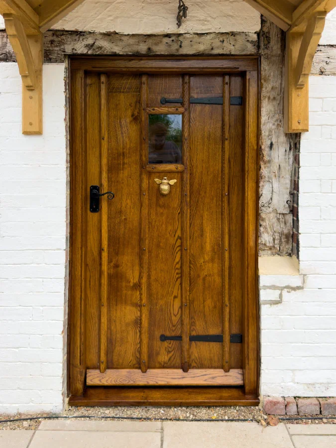 Oak front door with studded moulded ribs, a small glazed light, black strap hinges and a brass bee knocker, set in a timber-framed wall