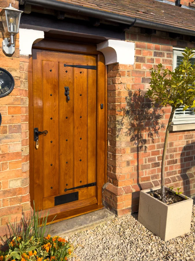 Studded oak front door with black strap hinges, a drop knocker and a letter plate in a brick porch