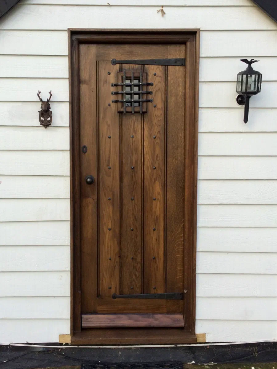 Dark-finished oak plank door with strap hinges and a small grille in a weatherboarded wall
