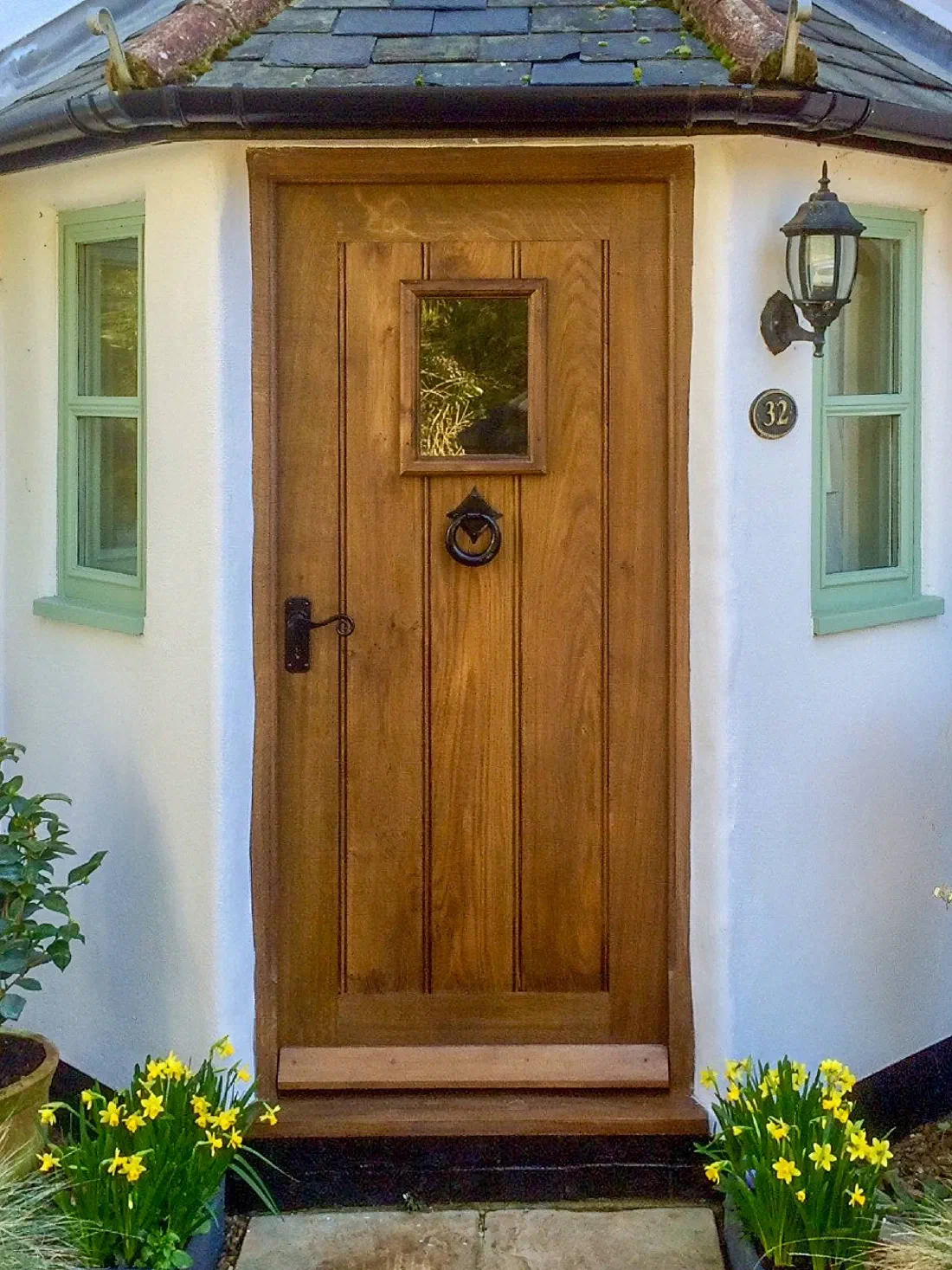 Framed oak plank door with a small glazed light, a ring knocker and a black lever handle, in a rendered cottage porch