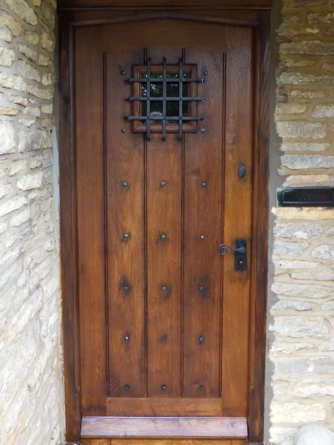 Oak framed plank front door with a Tudor-arched head rail, studwork, a small glazed light behind an iron grille and a lever handle, in a stone opening