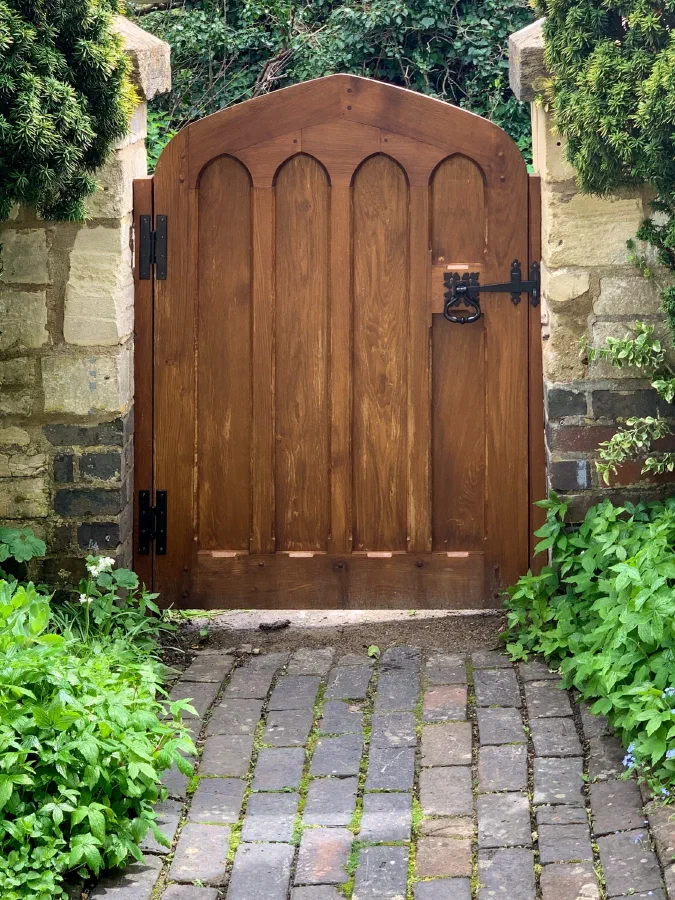 Arched oak garden door with pointed Gothic arches across its face and black strap hinges, in a stone wall